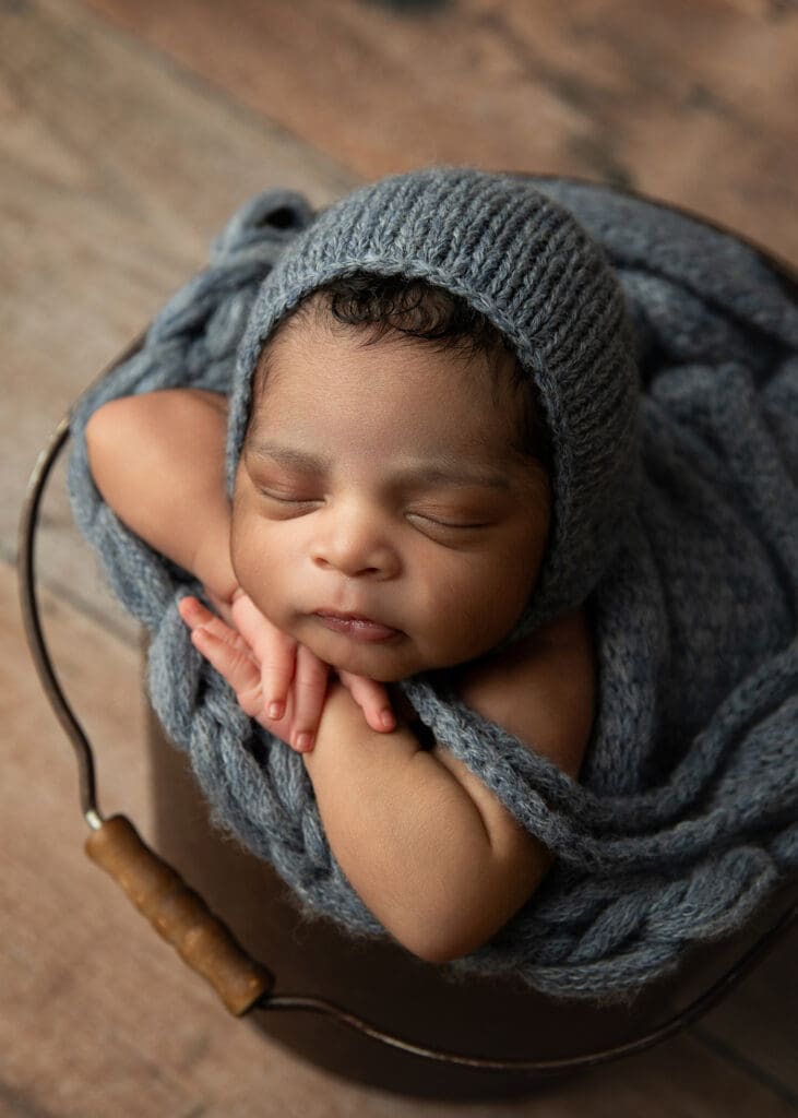 newborn baby boy with bonnet posed in bucket in Tulsa newborn studio